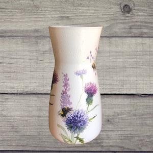 May include: White ceramic vase with a floral design featuring purple and blue flowers, green leaves, and a bumblebee.