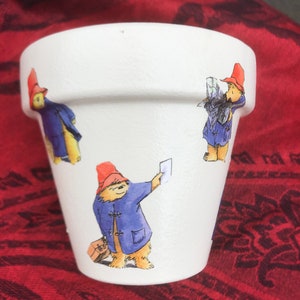 May include: White ceramic planter featuring a cartoon bear design. The bear is wearing a blue coat and a red hat. The planter has three different images of the bear.