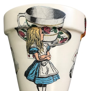 May include: White ceramic planter featuring a whimsical Alice in Wonderland design. The planter depicts a teacup and saucer with roses, a white rabbit, and Alice in a blue dress.