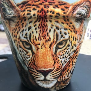 May include: A ceramic flower pot featuring a close-up image of a leopard's face. The pot is painted with a realistic image of a leopard's face, with brown, black, and white fur.