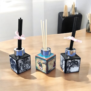 May include: Three decorative reed diffusers with square, illustrated bases. Each has a silver collar and a different arrangement of reeds or black sticks. One has a pink ribbon. The bases feature illustrations in black and white or colour.