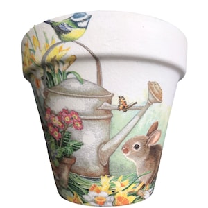 May include: A white ceramic flower pot with a spring-themed design. The pot features a watering can, a bluebird, a butterfly, and a rabbit. The design is in shades of green, yellow, pink, and brown.