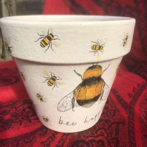 May include: White ceramic flower pot with a painted bee design and the text "bee happy".