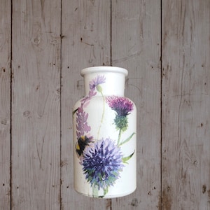 May include: White ceramic vase with a floral design featuring purple thistles and a bee. The vase has a round body and a narrow neck.