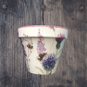 May include: A white ceramic flower pot with a pink rim, decorated with a floral pattern of purple flowers, green leaves, and bumblebees.
