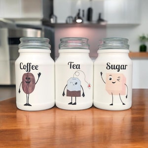 May include: Three white glass jars with glass lids, labelled "Coffee," "Tea," and "Sugar." Each jar features a cartoon illustration: a coffee bean, a teabag, and a sugar cube. The jars are on a wooden surface.