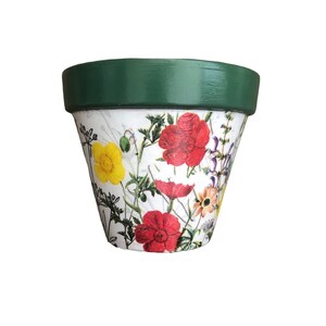 May include: A terracotta flower pot with a white base and a green rim. The pot is decorated with a floral design including red poppies, yellow buttercups, and other wildflowers. Perfect for planting flowers or herbs.