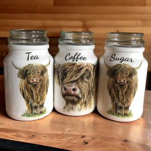 May include: Three white glass canisters with brown and white Highland cow illustrations. The canisters are labelled "Tea", "Coffee", and "Sugar".