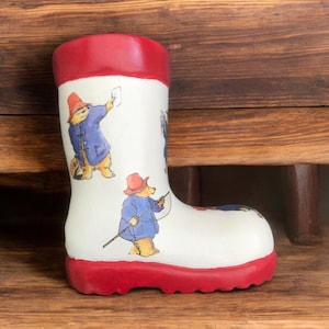 May include: A white ceramic boot with a red top and bottom. The boot is decorated with a repeating pattern of Paddington Bear wearing a blue coat and red hat.