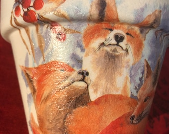 May include: White ceramic planter with a winter scene featuring a red robin and three foxes. The foxes are brown and orange with white accents. The background is a watercolor style with blue and purple hues.