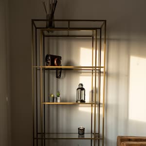 Könnte beinhalten: Ein goldfarbenes Regal mit Glasböden. Die Regale zeigen verschiedene dekorative Gegenstände, darunter eine Kamera, kleine Kakteen in Töpfen, eine Laterne, eine Kerze und einen Globus. Das Regal steht vor einer weißen Wand.