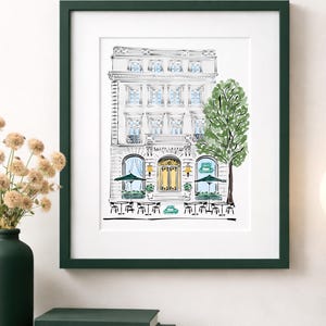 May include: A framed art print featuring a detailed sketch of a building facade with a restaurant. The artwork is in black and white with touches of color, including a green tree and umbrellas. The frame is dark green.