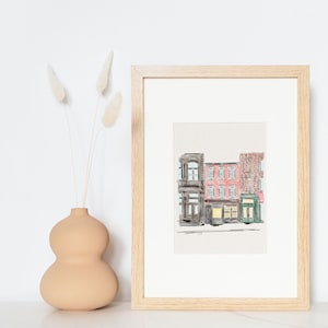 May include: A framed watercolor illustration of a row of three brick buildings with storefronts. The buildings are painted in shades of pink, brown, and green. The illustration is set against a white background.