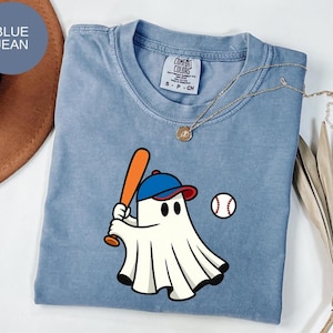 May include: A light blue Comfort Colors t-shirt featuring a cartoon ghost wearing a baseball cap and holding a bat. A baseball is next to the ghost. The shirt has a crew neck and short sleeves.