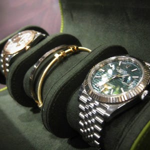 May include: A dark green watch case displays two watches and a bracelet. The watches have silver and gold elements, one with a green face. The bracelet is gold. The case is made of a soft, dark green material.