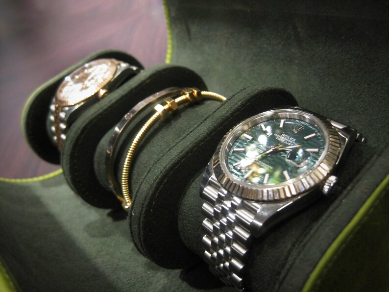 May include: A dark green watch case displays two watches and a bracelet. The watches have silver and gold elements, one with a green face. The bracelet is gold. The case is made of a soft, dark green material.