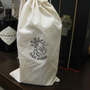 May include: A cream-coloured drawstring bag with a black graphic and text that reads "iskit atelye." The bag is standing on a dark brown surface. A bottle of gin and a black box are in the background.