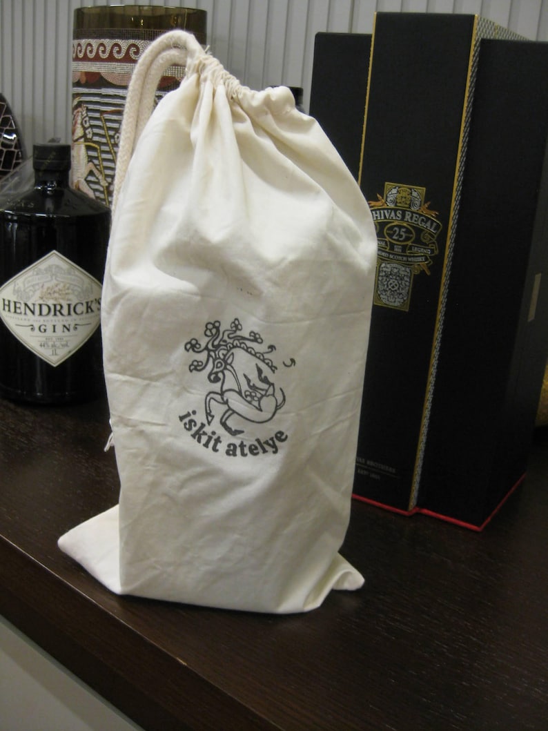 May include: A cream-coloured drawstring bag with a black graphic and text that reads "iskit atelye." The bag is standing on a dark brown surface. A bottle of gin and a black box are in the background.
