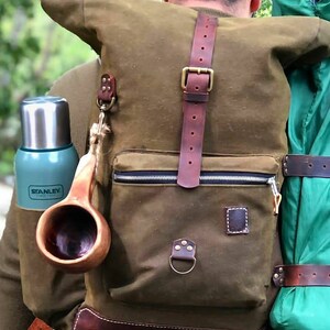 May include: A green canvas backpack with brown leather straps and buckles. The backpack has a zippered pocket and a small leather patch on the front. A wooden cup and a stainless steel water bottle with the word "STANLEY" are attached to the backpack.