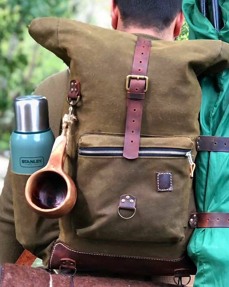 May include: A green canvas backpack with brown leather straps and buckles. The backpack has a zippered pocket and a small leather patch on the front. A wooden cup and a stainless steel water bottle with the word "STANLEY" are attached to the backpack.