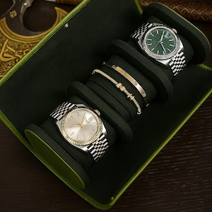 May include: A green watch case containing two silver and gold watches and two gold bracelets. The watches have silver and gold straps and light-coloured faces. The bracelets are gold with small decorative features. The case has a green trim.