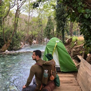 May include: A person sits on a wooden platform by a river, wearing a brown sweater and a large green and brown backpack. A green tent is pitched behind them.