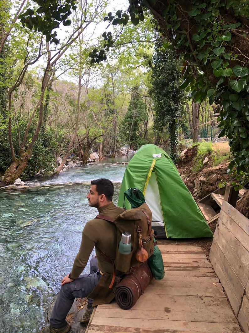 May include: A person sits on a wooden platform by a river, wearing a brown sweater and a large green and brown backpack. A green tent is pitched behind them.