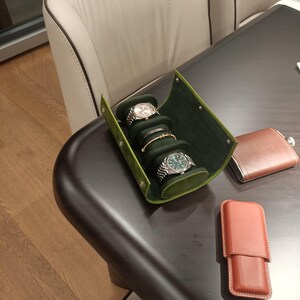 May include: A green leather watch roll displays three wristwatches and a bracelet. A brown leather flask and a brown leather case sit on a dark brown table. A beige chair is in the background.