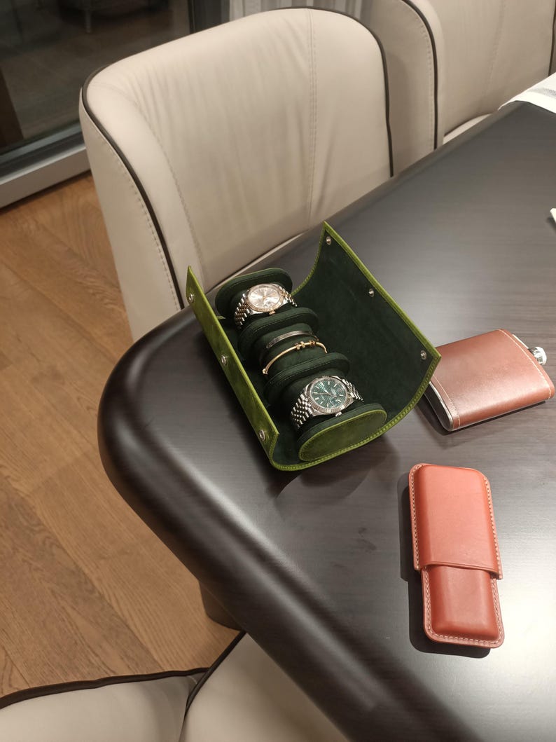 May include: A green leather watch roll displays three wristwatches and a bracelet. A brown leather flask and a brown leather case sit on a dark brown table. A beige chair is in the background.