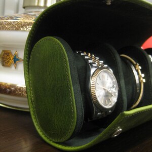 May include: A green leather watch travel case, open to reveal a silver and gold watch and a bracelet. The case has a black interior and a snap closure. The case is oval-shaped and made of green leather.