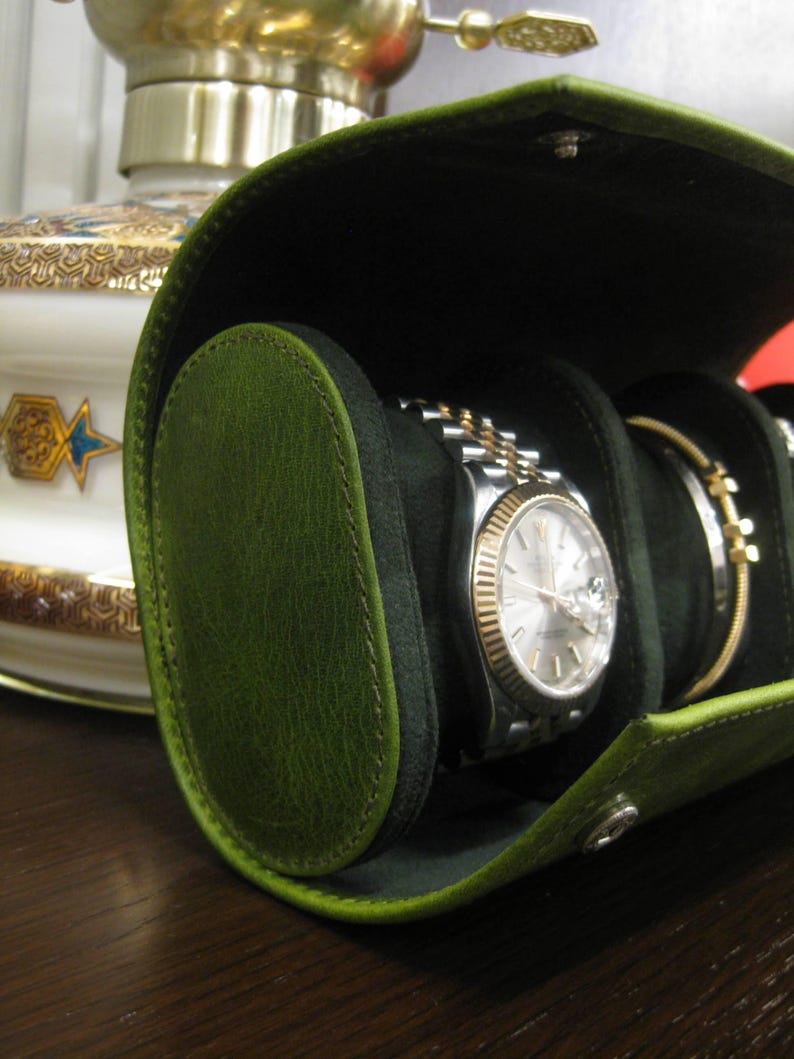 May include: A green leather watch travel case, open to reveal a silver and gold watch and a bracelet. The case has a black interior and a snap closure. The case is oval-shaped and made of green leather.