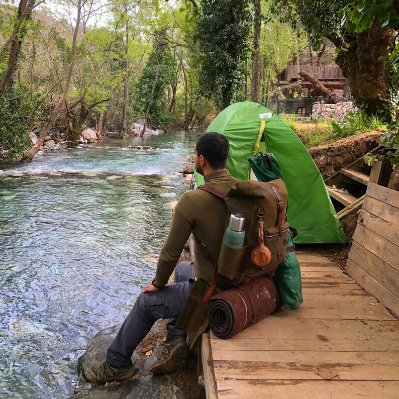 May include: A person sitting by a river with a green tent and a brown backpack. The backpack has a green water bottle and a wooden ornament attached to it.