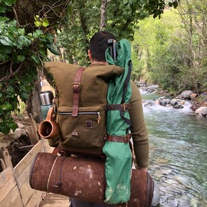 May include: A person wearing a brown and green backpack with leather straps and a rolled-up sleeping mat. The backpack is filled with camping gear, including a green tent, a thermos, and a mug. The person is walking on a wooden bridge over a stream.