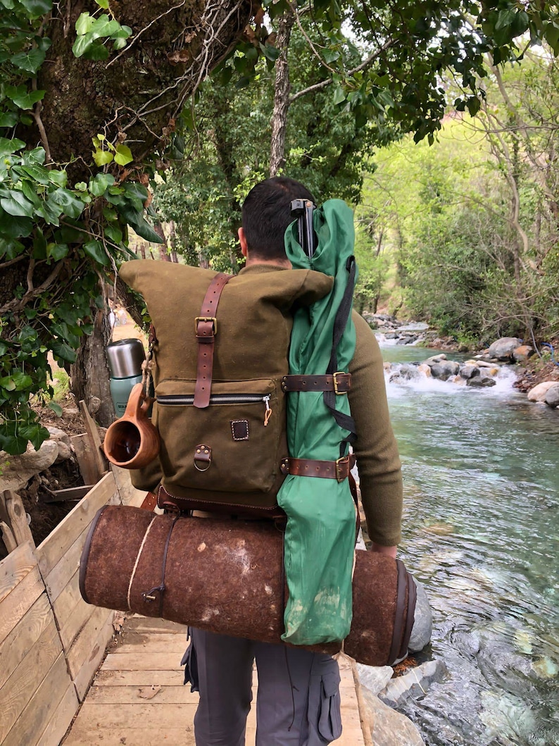 May include: A person wearing a brown and green backpack with leather straps and a rolled-up sleeping mat. The backpack is filled with camping gear, including a green tent, a thermos, and a mug. The person is walking on a wooden bridge over a stream.