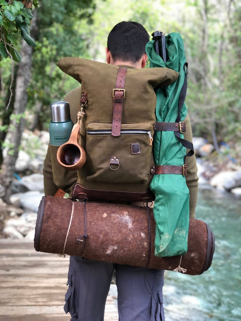 May include: A person wearing a green and brown canvas backpack with leather straps and buckles. The backpack is carrying a green sleeping bag, a brown rolled-up sleeping mat, a green thermos, and a wooden cup.