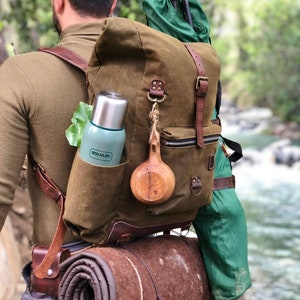 May include: A person wearing a brown canvas backpack with leather straps and buckles. The backpack has a green insulated water bottle, a wooden cup, and a rolled-up sleeping mat attached to it.