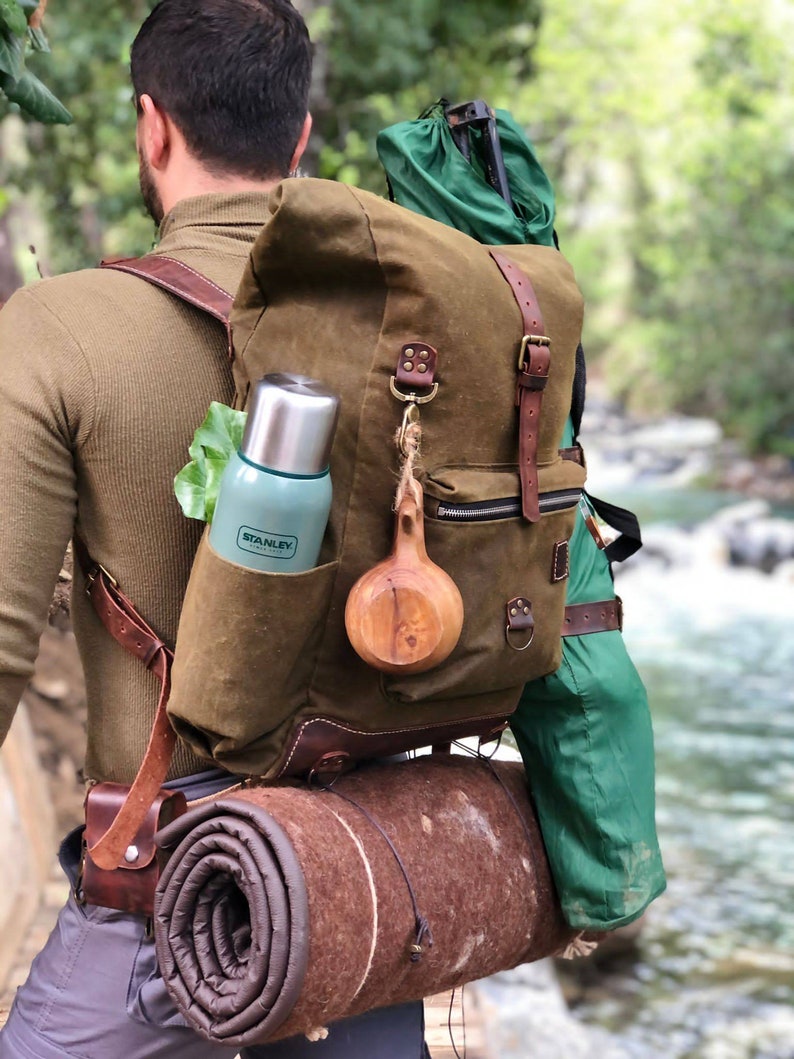 May include: A person wearing a brown canvas backpack with leather straps and buckles. The backpack has a green insulated water bottle, a wooden cup, and a rolled-up sleeping mat attached to it.