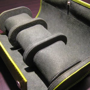 May include: Open, grey watch case with three padded compartments. The interior is lined with a soft, dark grey material. The case is trimmed with a bright yellow-green border and has a snap closure.
