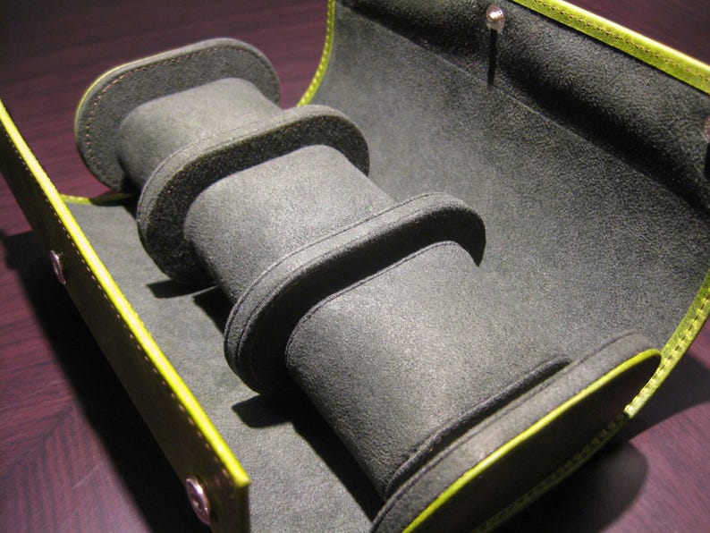 May include: Open, grey watch case with three padded compartments. The interior is lined with a soft, dark grey material. The case is trimmed with a bright yellow-green border and has a snap closure.
