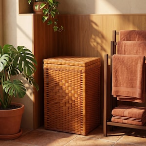 Puede incluir: Un cesto para la ropa rectangular fabricado con mimbre trenzado de color marrón claro, con tapa plana y un diseño vertical estructurado. El cesto está situado en una habitación con baldosas de terracota y paneles de madera en la pared.