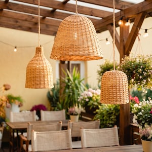 Könnte beinhalten: Drei gewebte Pendelleuchten aus Rattan in verschiedenen Formen hängen von einer Holzpergola. Die Leuchten haben eine warme, natürliche, hellbraune Farbe und werfen ein sanftes Licht. Die Szene spielt sich im Freien mit einem Esstisch und Stühlen ab.