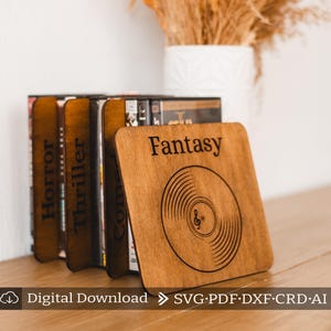 Puede incluir: Separadores de libros de madera con un diseño de disco y la palabra "Fantasy" grabada en la parte delantera. Los separadores de libros son de color marrón y tienen un acabado de madera natural. Los separadores de libros se utilizan para sostener una pila de DVD.
