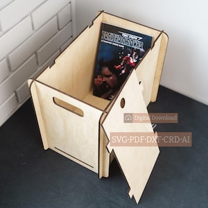 Puede incluir: Una caja de almacenamiento de madera de color claro con un libro dentro. La caja tiene forma rectangular con un asa recortada en un lado y una puerta con bisagras. El libro tiene una cubierta oscura con texto en idioma extranjero.