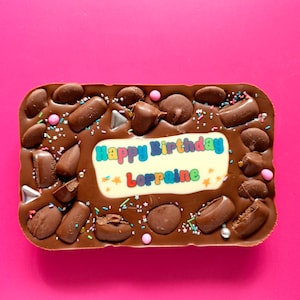 May include: A chocolate bar with a white background and colourful text that says "Happy Birthday Lorraine". The chocolate bar is covered in chocolate chips, sprinkles, and Cadbury chocolate pieces.