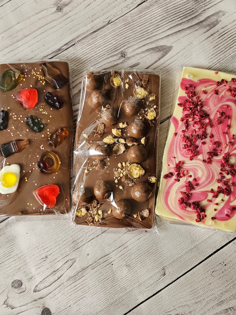 May include: Three different chocolate bars wrapped in plastic. The first bar is milk chocolate with sprinkles, gummy candies, and a fried egg. The second bar is milk chocolate with chocolate chips and peanuts. The third bar is white chocolate with swirls of pink and red and red sprinkles.