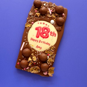 May include: A chocolate bar with a white chocolate design that says "18th Happy Birthday Amy" with red sprinkles and chocolate sweets.