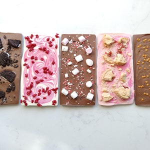 May include: Four different chocolate bars with various toppings. The bars are arranged horizontally from left to right. The first bar is dark chocolate with Oreo cookie pieces. The second bar is white chocolate with pink sprinkles. The third bar is milk chocolate with marshmallows and sprinkles. The fourth bar is pink chocolate with shortbread cookie pieces.