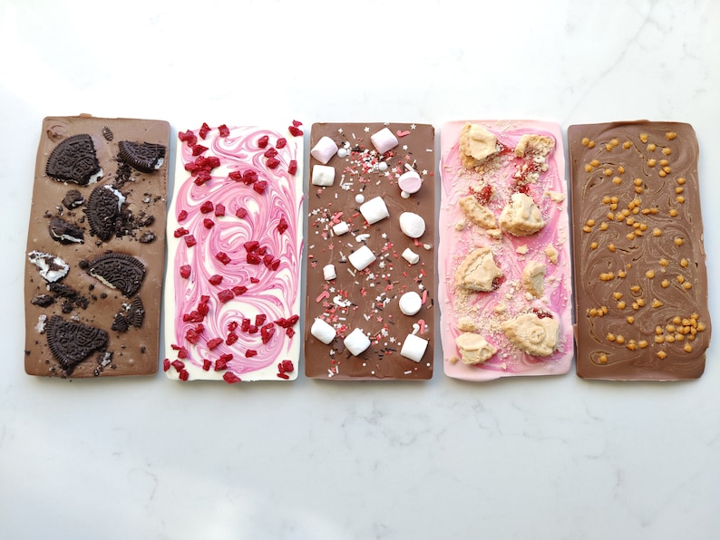 May include: Four different chocolate bars with various toppings. The bars are arranged horizontally from left to right. The first bar is dark chocolate with Oreo cookie pieces. The second bar is white chocolate with pink sprinkles. The third bar is milk chocolate with marshmallows and sprinkles. The fourth bar is pink chocolate with shortbread cookie pieces.