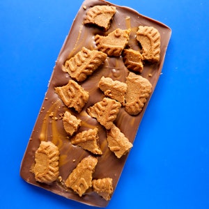 May include: A chocolate bar with broken pieces of shortbread cookies on top. The chocolate is a light brown color and the cookies are a golden brown color. The bar is on a blue background.