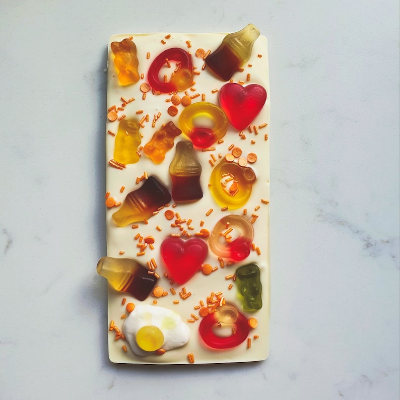 May include: A white chocolate bar with a variety of colorful gummy candies, including hearts, rings, and soda bottles. The bar is decorated with gold sprinkles.